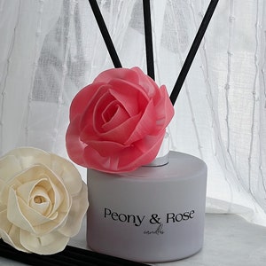 Könnte beinhalten: Eine weiße Diffuserflasche mit schwarzen Schilfrohren und einer rosa Rose obenauf. Die Flasche trägt den Schriftzug "Peony & Rose candles" auf der Vorderseite.