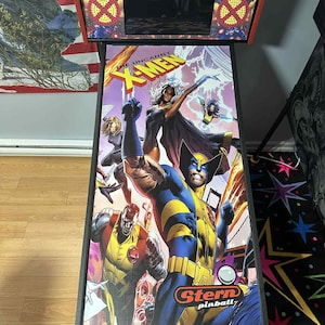May include: An arcade pinball machine with a colourful comic book design of X-Men characters. The artwork includes Wolverine, Storm, and other heroes. The machine has a black base and a red top with the text "THE UNCANNY X-MEN" and "Stern pinball".
