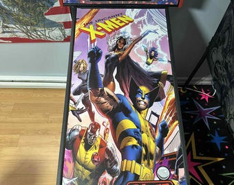 May include: An arcade pinball machine featuring a vibrant comic book illustration of X-Men characters. The artwork includes Wolverine, Storm, and other heroes. The machine has a black base and a red top with the text "THE UNCANNY X-MEN" and "Stern pinball".