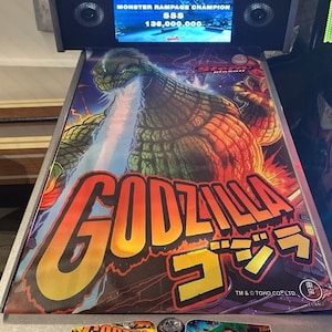 Puede incluir: Una máquina de pinball con temática de Godzilla. El campo de juego presenta una gran ilustración de Godzilla lanzando fuego, con el título del juego en letras naranjas y amarillas en negrita. La pantalla de la máquina muestra una puntuación alta de 136.000.000.