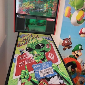 Puede incluir: Un extraterrestre verde con bigote y un cartel que dice "Normal Human Seeking Transport to Isleta Amphitheater Albuquerque, NM" aparece en un campo de juego de una máquina de pinball. El campo de juego es verde y azul con un logotipo de Stern Pinball. La máquina se titula "Foo Fighters".