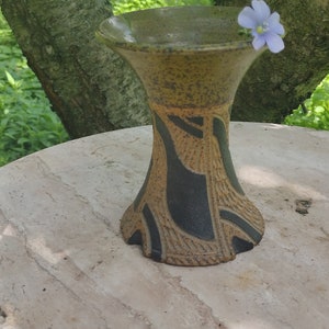 Peut inclure: Un petit vase en céramique brun avec des motifs géométriques noirs et bruns. Le vase est posé sur une surface brun clair avec deux fleurs bleu clair.