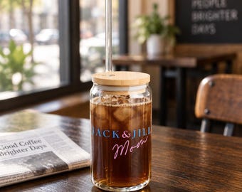 May include: A clear glass tumbler with a bamboo lid and a glass straw, filled with dark iced coffee. The glass features the blue text "JACK & JILL" above the pink cursive text "Mom" printed on the front surface of the cylindrical container.