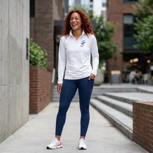 May include: A white long-sleeve quarter-zip pullover with a small colorful graphic on the chest, paired with solid navy blue leggings featuring side pockets and white athletic sneakers with pink accents on the soles and heel tabs.