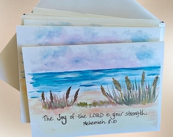 May include: A stack of rectangular greeting cards featuring a watercolor painting of a beach scene with blue ocean waves, sandy dunes, and tall grasses. The text "The Joy of the LORD is your strength. Nehemiah 8:10" is written in black ink.
