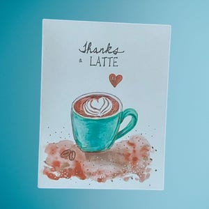 May include: A watercolour greeting card featuring a teal coffee cup with a latte and heart-shaped foam art. The words "Thanks a LATTE" are written above the cup, with a small red heart. Coffee beans and brown watercolour accents are at the bottom.