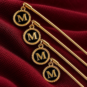 May include: Four gold-toned metal hair pins, each featuring a circular charm with a black enamel center and a gold letter M. The charms have a beaded edge and are attached to the long, slender, straight gold pins by a small jump ring.