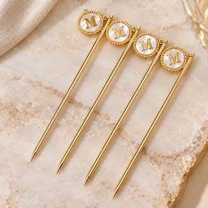 May include: Four gold-toned metal cocktail picks, each featuring a circular charm at the top. The charm has a beaded gold border, a white mother-of-pearl inlay, and a gold-colored letter M centered on the surface of the inlay.