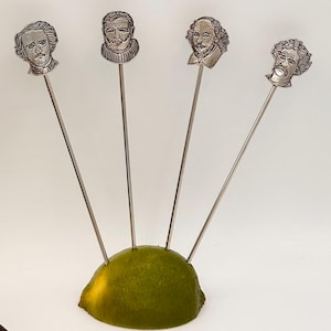 Può includere: Quattro stuzzicadenti d'argento con ritratti incisi di figure letterarie, tra cui Edgar Allan Poe e William Shakespeare, sono inseriti in una metà di lime verde. Gli stuzzicadenti hanno steli lunghi e sottili e sono disposti a ventaglio.
