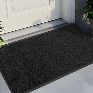May include: Black doormat with a dark grey border. The doormat is made of a textured material and is placed in front of a white door.