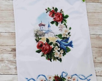 May include: White embroidered fabric with a blue bird, red roses, and white flowers surrounding a church building. The fabric has a white lace border.
