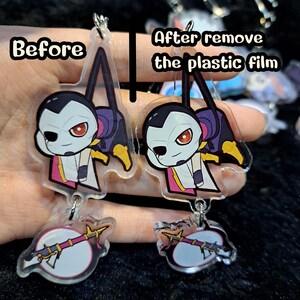 May include: Two clear acrylic charms with a cartoon character design. The charm on the left has a protective plastic film on it, while the charm on the right has the film removed. Both charms feature a character with black hair, a white face, and red eyes. The character is holding a weapon in their right hand. The charms are attached to a silver chain.