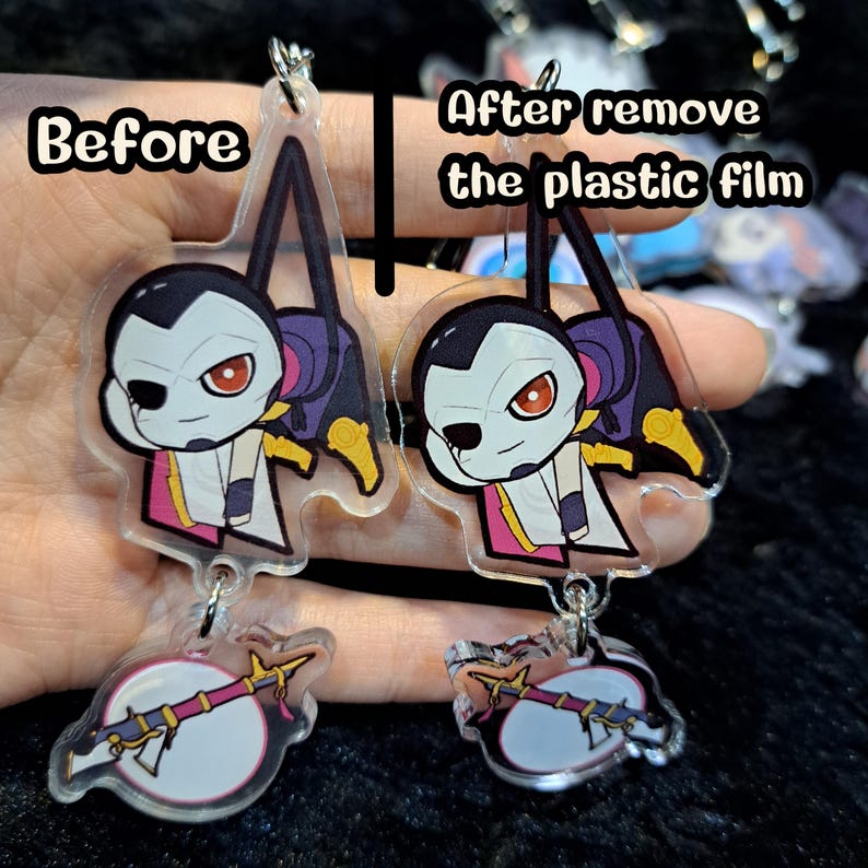 May include: Two clear acrylic charms with a cartoon character design. The charm on the left has a protective plastic film on it, while the charm on the right has the film removed. Both charms feature a character with black hair, a white face, and red eyes. The character is holding a weapon in their right hand. The charms are attached to a silver chain.