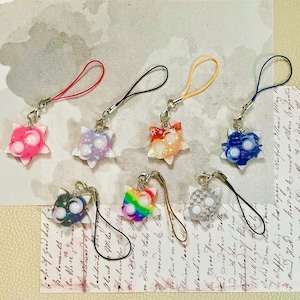 Può includere: Sette ciondoli a forma di stella con cordini per l'aggancio. I ciondoli presentano vari colori tra cui rosa, viola, arancione, blu, verde scuro, arcobaleno e grigio, ognuno con due accenti circolari bianchi e piccoli glitter a forma di stella.
