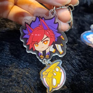 May include: A clear acrylic keychain featuring a cartoon character with red hair, purple and blue hair, and a yellow and white outfit. The character is holding a sword. The keychain also features a separate charm of a yellow and white dragon head.