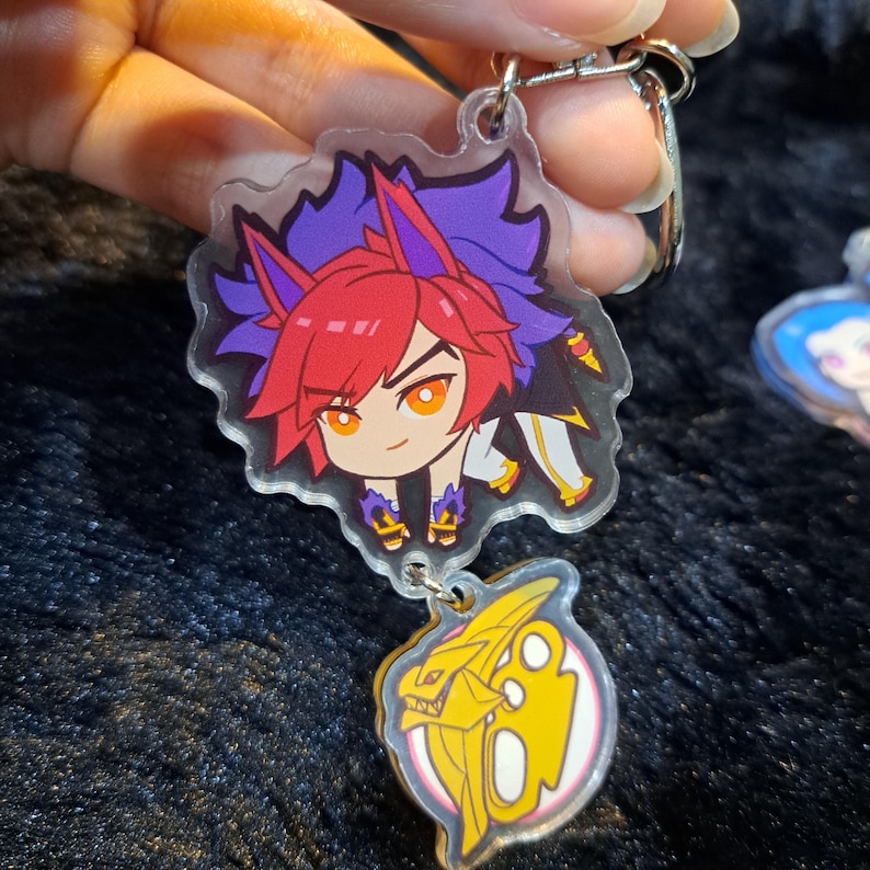 May include: A clear acrylic keychain featuring a cartoon character with red hair, purple and blue hair, and a yellow and white outfit. The character is holding a sword. The keychain also features a separate charm of a yellow and white dragon head.