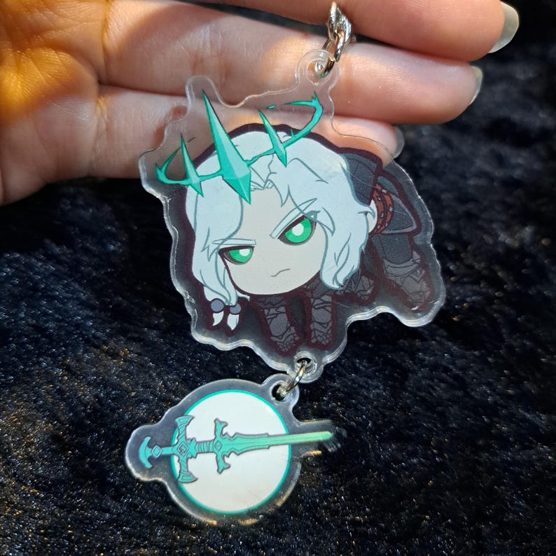 May include: A clear acrylic keychain featuring a cartoon character with white hair, a green crown, and a green sword. The character is wearing a black and white outfit and is holding a sword.
