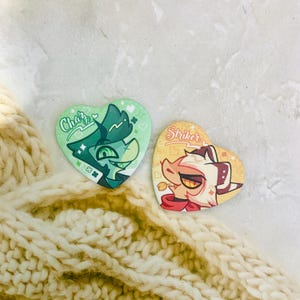 May include: Two heart-shaped pin-back buttons featuring cartoon character illustrations. The left button is green with the text Chaz, and the right button is yellow with the text Striker. Both designs include small heart and star accents.