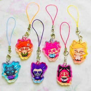 Puede incluir: Seis dijes de acrílico con personajes de dibujos animados estilizados con orejas de gato, cada uno unido a un cordón de bucle de colores. Los personajes muestran varios atuendos y accesorios, incluyendo máscaras, sombreros y gafas, en colores vibrantes.