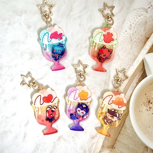 Könnte beinhalten: Sechs bunte Acryl-Schlüsselanhänger in Form von Eisbechern, jeder mit einem anderen Cartoon-Charakter. Die Schlüsselanhänger haben goldfarbene sternförmige Verschlüsse. Die Eisbecher haben rosa, blaue, grüne und gelbe Akzente.