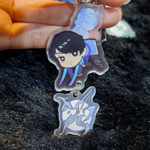 May include: A clear acrylic keychain with two charms. The top charm features a cartoon character with black hair, blue eyes, and a blue and purple outfit. The bottom charm features a stylized design of a sword and a circle with a white background.