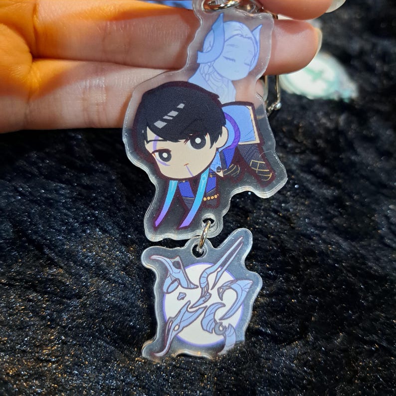 May include: A clear acrylic keychain with two charms. The top charm features a cartoon character with black hair, blue eyes, and a blue and purple outfit. The bottom charm features a stylized design of a sword and a circle with a white background.