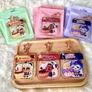 May include: Three clear plastic keychains with colourful cartoon characters and text. The keychains are on a wooden surface. The text on the keychains reads "Strainer", "Blitz", and "Scolus".