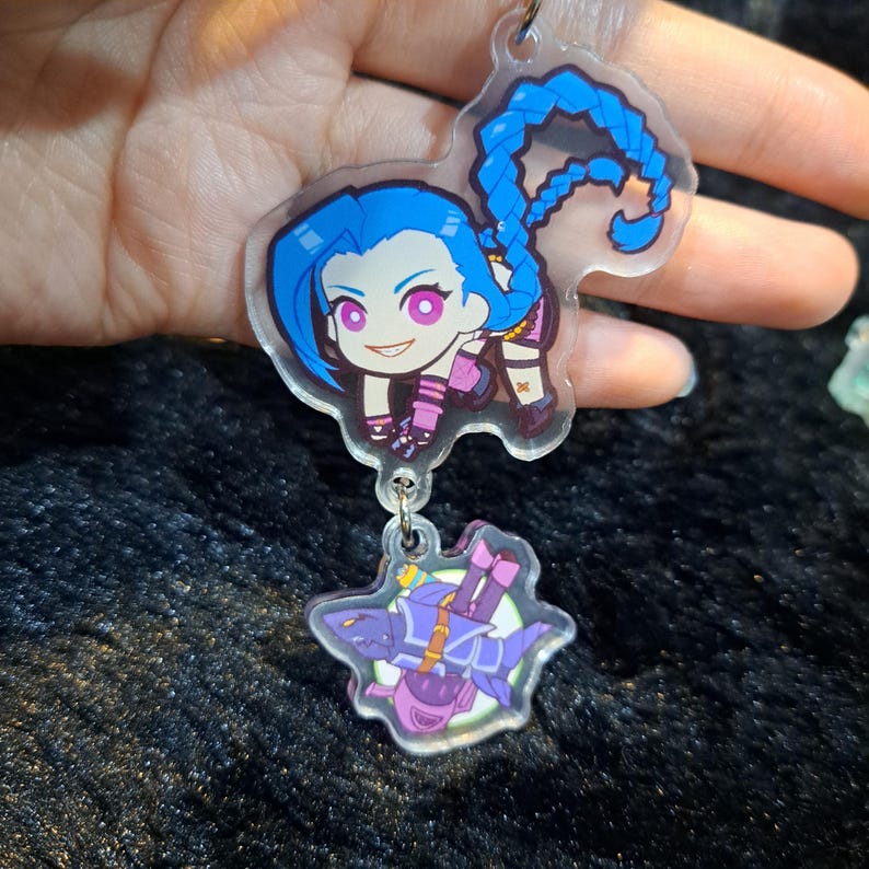 May include: A clear acrylic keychain featuring a cartoon character with blue hair and a purple outfit. The character is holding a weapon and is attached to a smaller charm of a purple creature.