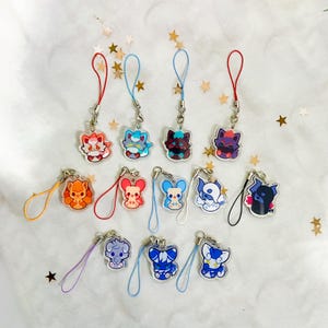 Puede incluir: Una colección de doce dijes de acrílico con criaturas de dibujos animados estilizadas en varios colores, incluyendo naranja, azul, morado y negro. Cada dije está unido a un lazo de cordón de color y presenta un diseño de personaje único.