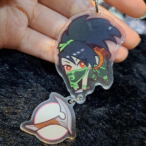 May include: A clear acrylic keychain featuring a cartoon character with black hair, a green mask, and a green and orange outfit. The character is holding a weapon. The keychain also includes a separate clear acrylic charm with a white and pink design.