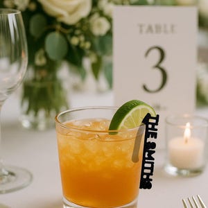 May include: A cocktail in a clear glass with a lime wedge and a black name tag that reads "The Smiths." The drink has an orange hue and is garnished with a lime slice. A table number 3 card and a lit candle are in the background.