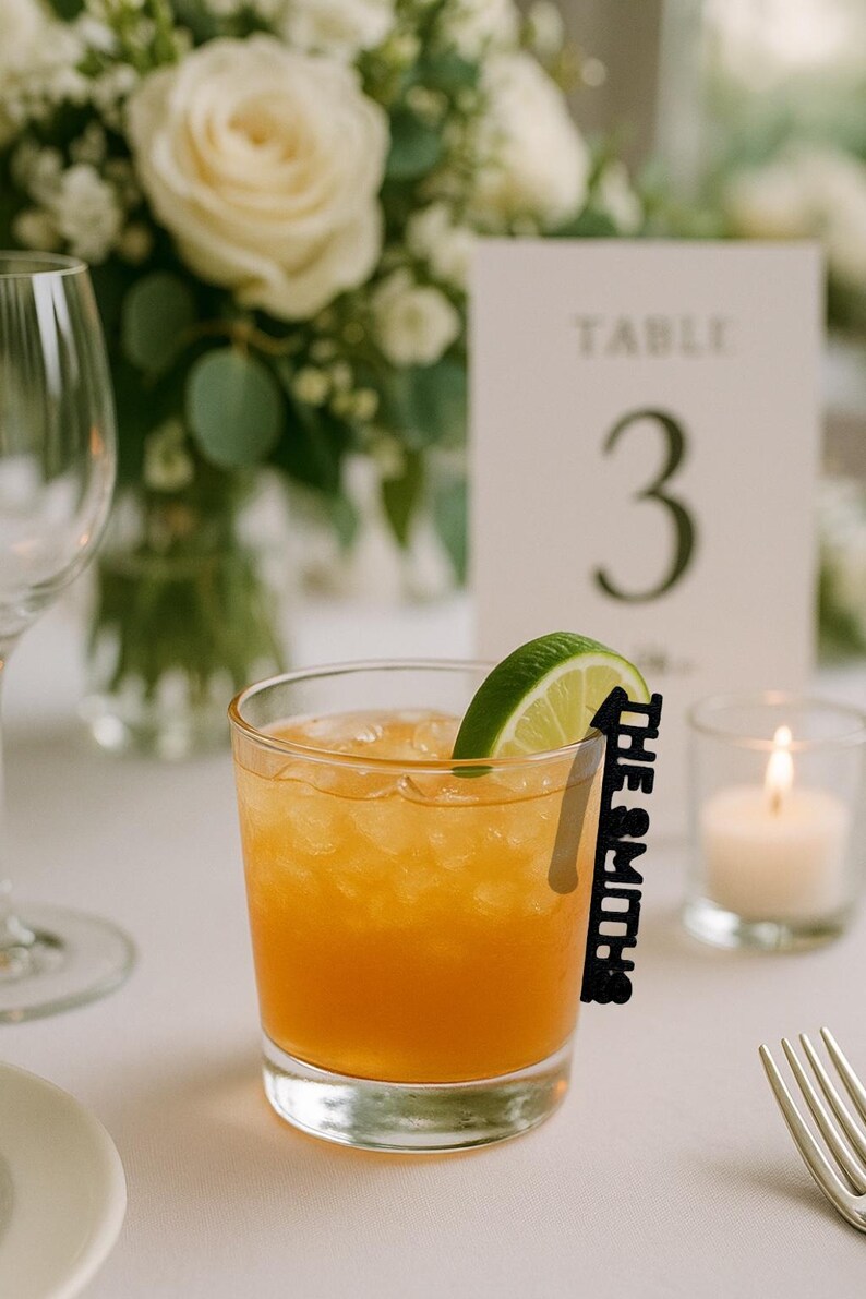 May include: A cocktail in a clear glass with a lime wedge and a black name tag that reads "The Smiths." The drink has an orange hue and is garnished with a lime slice. A table number 3 card and a lit candle are in the background.