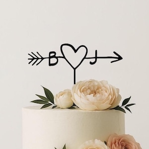 May include: Black metal cake topper featuring a heart and arrow design with the letters "B" and "J". The topper is placed on a white cake decorated with cream and peach-colored flowers and green leaves. A wedding cake decoration.