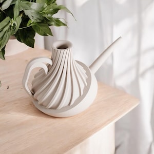 May include: A white ceramic watering can with a unique, folded design and a long spout. The can has a handle and is sitting on a light wood surface.