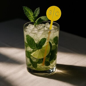 Op de afbeelding: Een verfrissende mojito cocktail in een helder glas. De drank is gevuld met ijs, muntblaadjes en een limoenpartje. Een gele drankroerder met de letters "S/C" is in de drank geplaatst. De drank staat op een witte ondergrond.