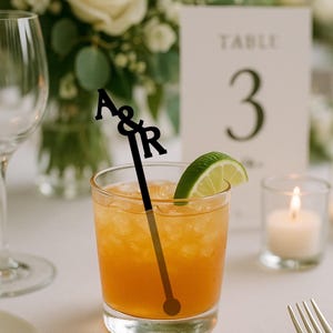 Op de afbeelding: Een cocktail met een partje limoen en een zwarte roerstaaf met de letters "A&R" bovenaan. De drank zit in een helder glas en een brandende kaars en tafelnummer 3 staan op de achtergrond. De drank is oranje.