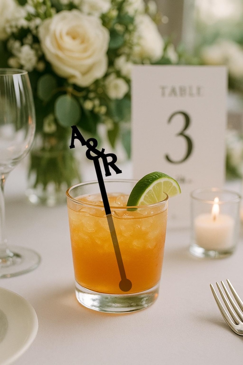 Op de afbeelding: Een cocktail met een partje limoen en een zwarte roerstaaf met de letters "A&R" bovenaan. De drank zit in een helder glas en een brandende kaars en tafelnummer 3 staan op de achtergrond. De drank is oranje.