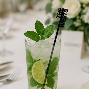 Op de afbeelding: Een verfrissende mojito-cocktail in een helder glas, gegarneerd met muntblaadjes en een schijfje limoen. Een zwarte roerstaaf met de tekst "THE WESTS" zit in de drank. De drank staat op een witte tafel.