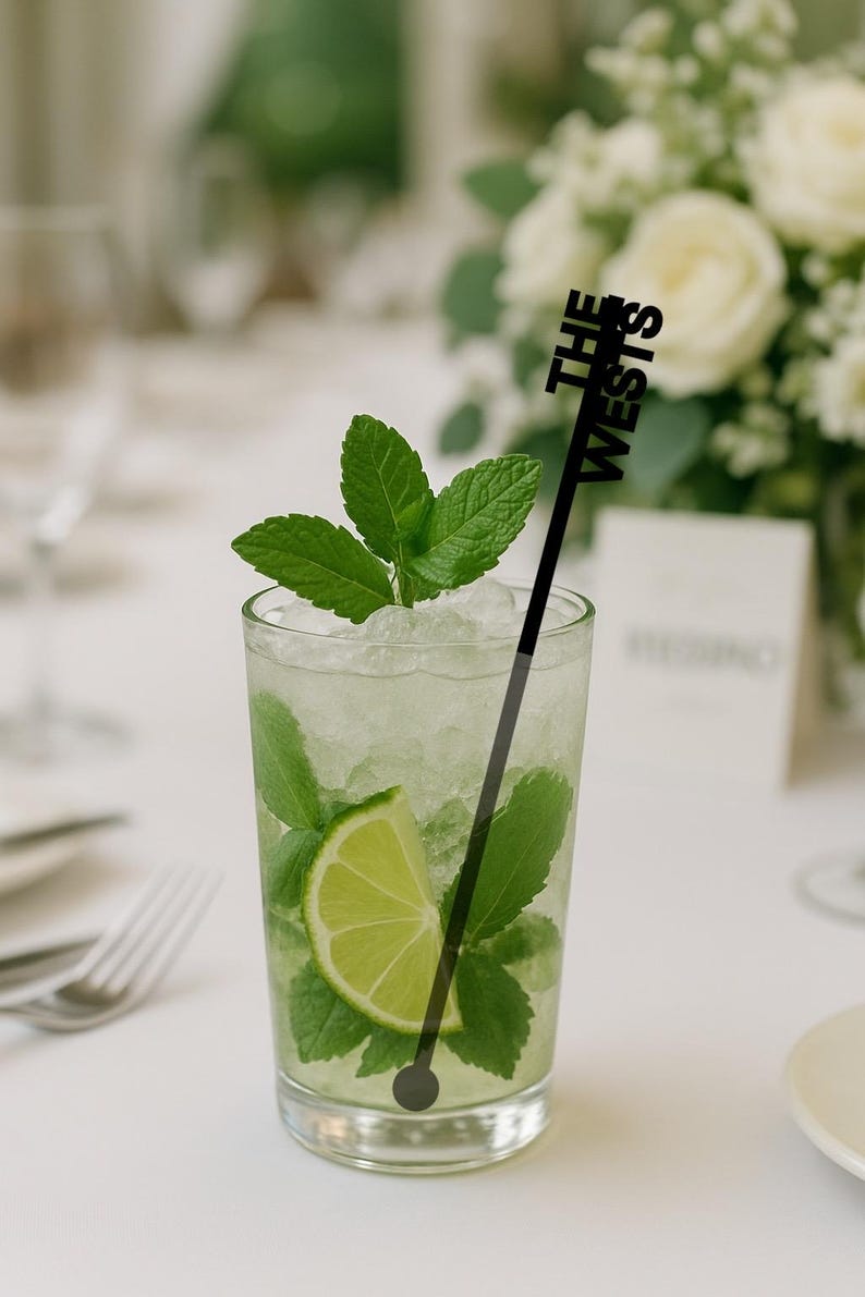 Op de afbeelding: Een verfrissende mojito-cocktail in een helder glas, gegarneerd met muntblaadjes en een schijfje limoen. Een zwarte roerstaaf met de tekst "THE WESTS" zit in de drank. De drank staat op een witte tafel.