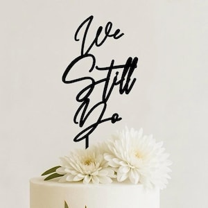 May include: A black cake topper with the words "We Still Do" in elegant script. The topper is placed on a white cake, adorned with white flowers and green leaves. A wedding or anniversary theme.