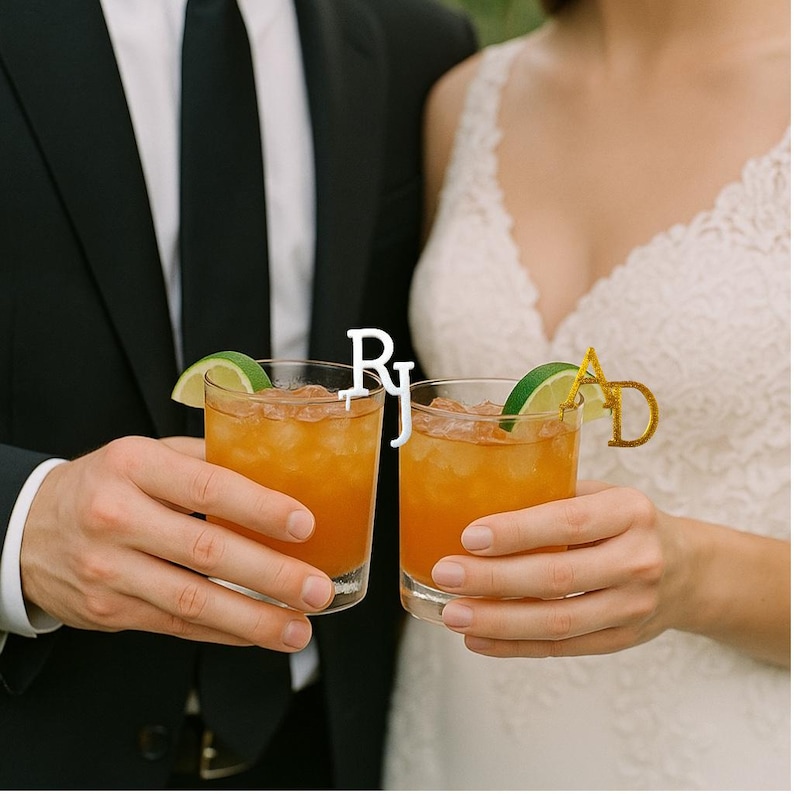 May include: Two glasses of an orange-colored cocktail with ice and lime wedges. One glass has a gold letter "AD" and the other has the letters "RJ". The image shows a person in a suit and a person in a wedding dress holding the glasses.