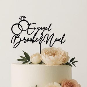 May include: A black cake topper with the words "Engaged Brooke & Noah" in cursive script, featuring a diamond ring and two interlocking rings. The topper is placed on a white cake decorated with flowers and greenery.