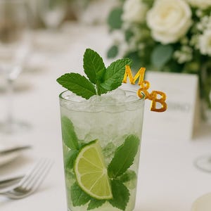May include: A refreshing mojito cocktail in a tall glass, garnished with fresh mint leaves and a lime wedge. The drink is topped with crushed ice and a gold-colored '&' and initials 'M&B' decoration. The background includes a white table setting and a bouquet of white flowers.