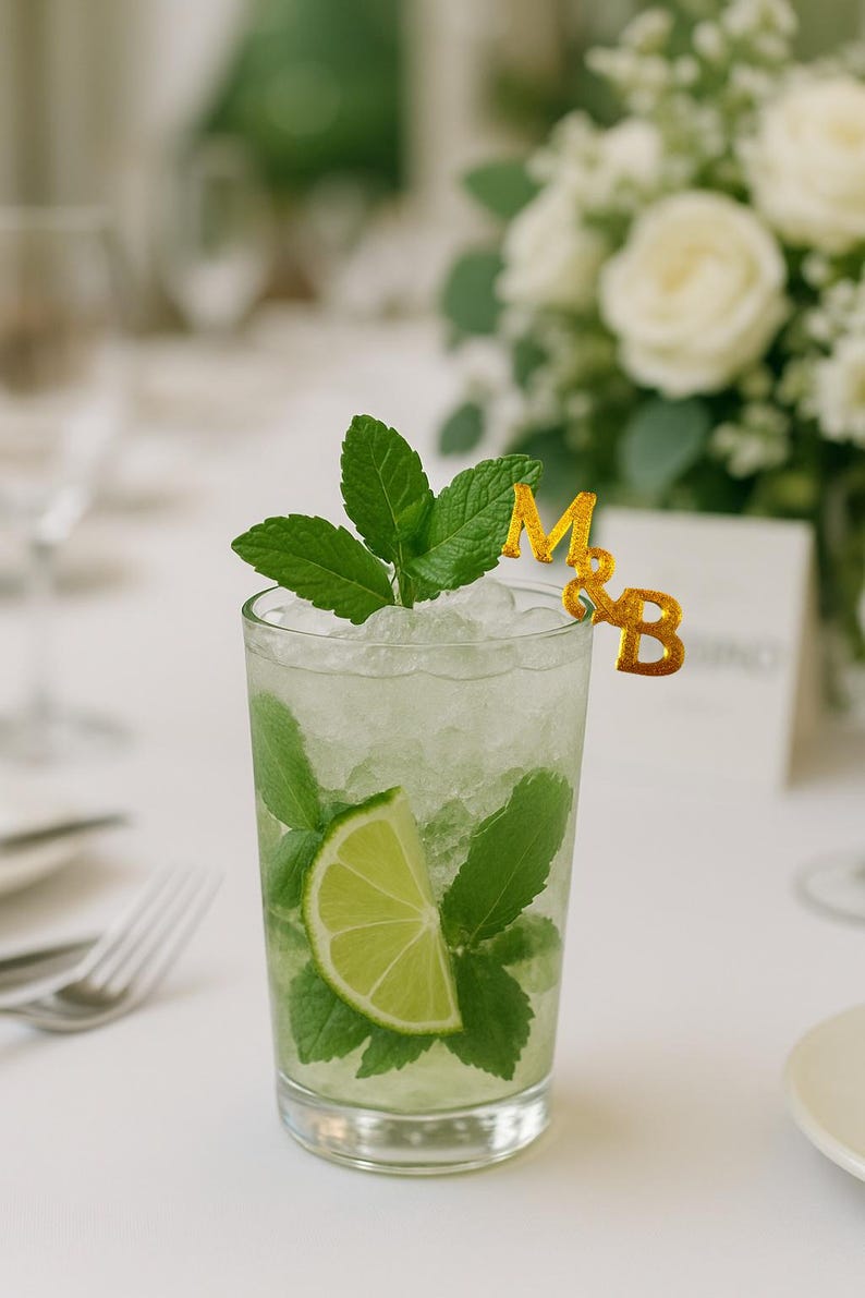 May include: A refreshing mojito cocktail in a tall glass, garnished with fresh mint leaves and a lime wedge. The drink is topped with crushed ice and a gold-colored '&' and initials 'M&B' decoration. The background includes a white table setting and a bouquet of white flowers.