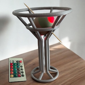 May include: A gray 3D printed martini glass with a round base and a sphere inside. The sphere is lit with a red light and is held in place by a wooden skewer. A remote control with buttons for different colored lights is in the foreground.