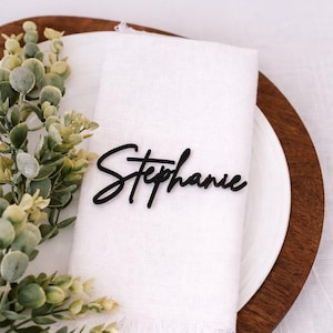 May include: A black acrylic nameplate with the name "Stephanie" in a cursive font rests on a white linen napkin. The napkin is placed on a white plate, which is set on a wooden charger. Greenery accents the scene.