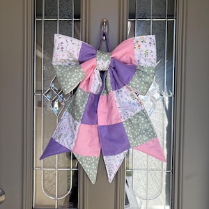 May include: A large, decorative bow made of fabric, featuring a patchwork design in shades of pink, purple, and green. The bow is adorned with floral patterns and hangs from a metal hook, suitable for door decor.