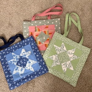 Puede incluir: Tres bolsas de mano hechas a mano con diseños de estrellas acolchadas. Una bolsa es azul con estrellas florales blancas, otra es verde con estrellas florales blancas, y la tercera es rosa y naranja con un estampado floral. Cada bolsa tiene un asa de diferente color.