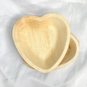 May include: Two heart-shaped, disposable plates made from natural, light-brown palm leaf material. The plates have a textured surface with visible grain and are stacked on a white background. The plates are ideal for serving appetizers or desserts.