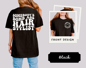 Może przedstawiać: Czarny t-shirt z białym napisem "SOMEBODY'S BOMB ASS HAIR STYLIST" z tyłu. Przedni wzór przedstawia uśmiechniętą buzię z napisem "BOMB ASS HAIR STYLIST". Koszulka ma swobodny styl.